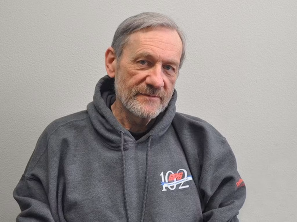 Michael Bronski seated against a plain light gray wall, wearing a dark gray hoodie with the logo "GFS 102" on it. He has a trimmed gray beard and a soft, thoughtful expression.