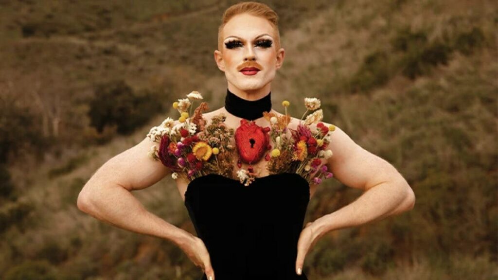 Pattie Gonia stands confidently outdoors wearing a black strapless dress adorned with vibrant dried flowers across the chest. The background is blurred forest terrain.