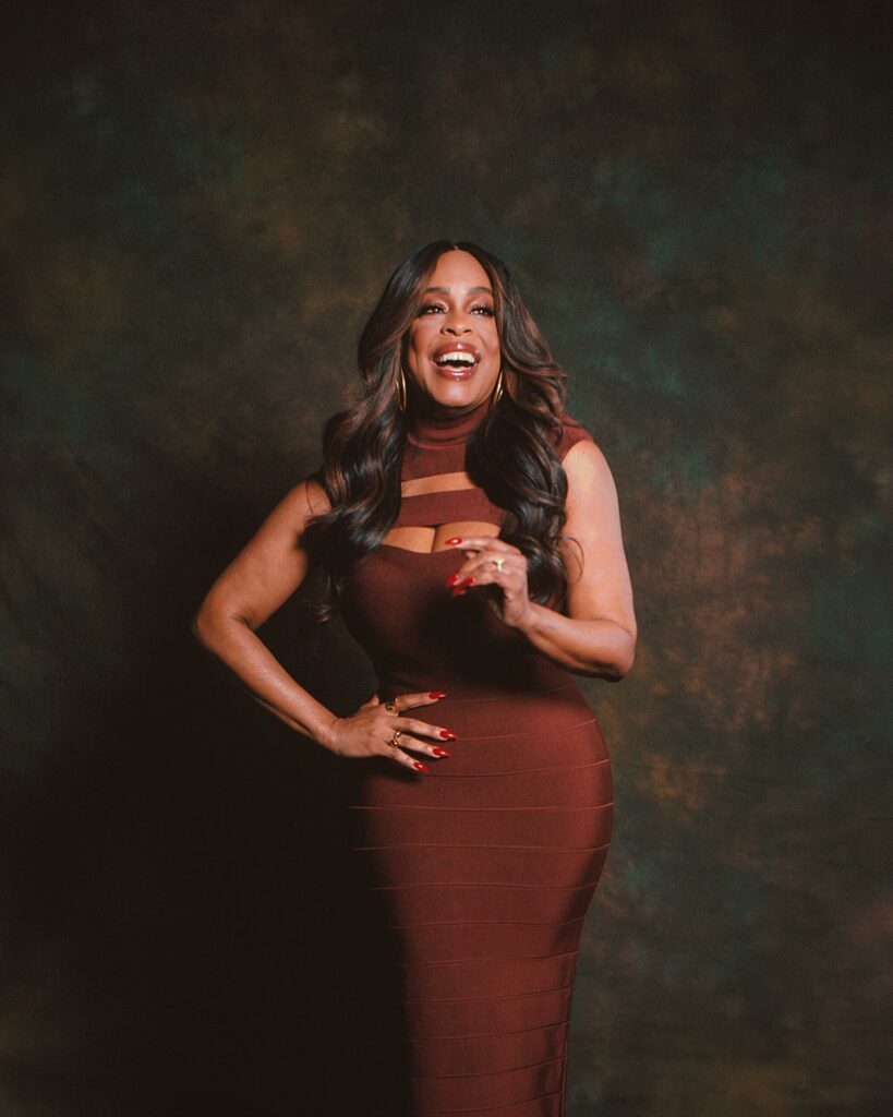 Niecy Nash-Betts beaming with laughter in a burgundy bodycon dress with cutout details, standing in front of a dark mottled backdrop with her hands mid-gesture.