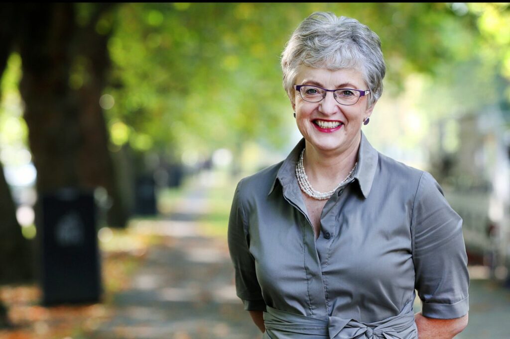 Katherine Zappone standing on a leafy walkway wearing an olive-colored dress and pearls, smiling with soft sunlight in the background.