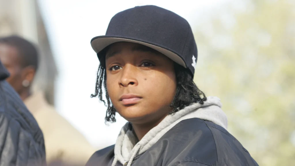 Felicia Pearson in character as Snoop from The Wire, wearing a black flat-brim hat, black jacket, and gray hoodie. She appears youthful and stoic, with soft lighting and blurred figures in the background.