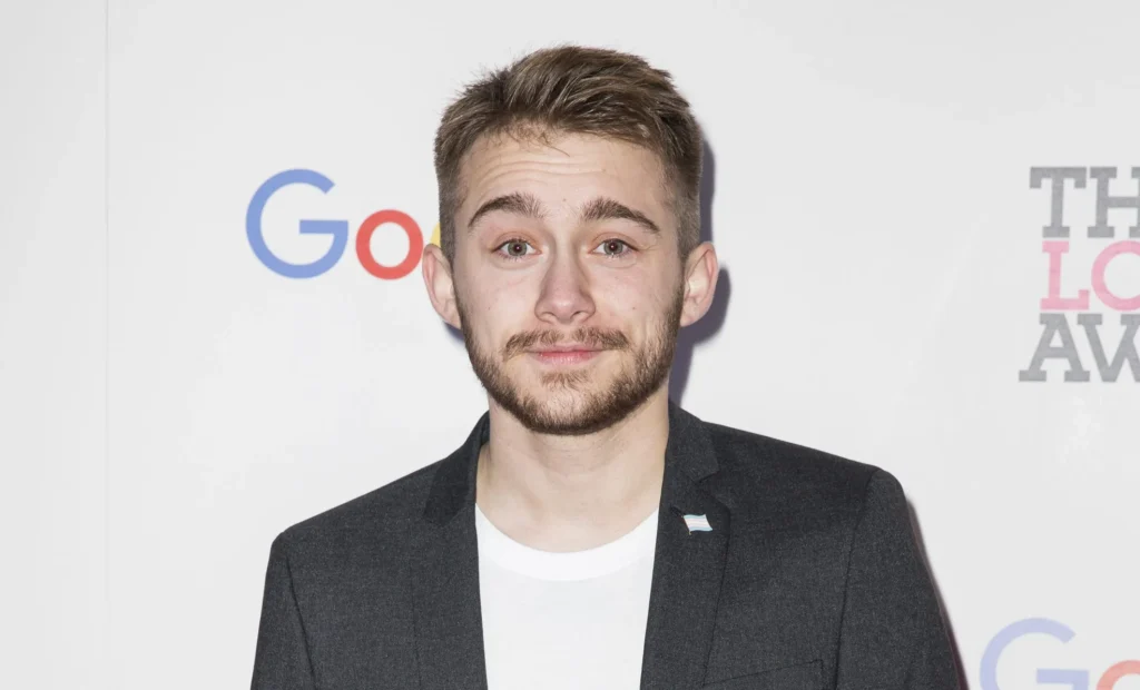 Alex Bertie at a formal event, wearing a black blazer over a white shirt. A small trans pride flag pin is on his lapel. He has a trimmed beard and is smiling with a white background featuring logos for Google and The Lovie Awards.