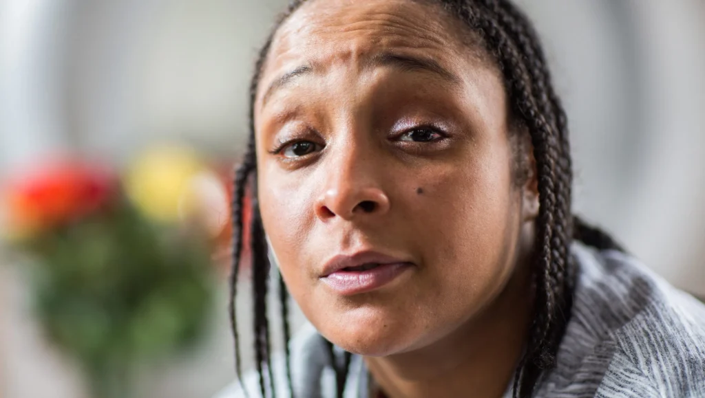 Close-up shot of Felicia Pearson indoors, showing detailed facial texture and braids. Her expression is pensive and direct. Soft-focus colored flowers are visible in the background.