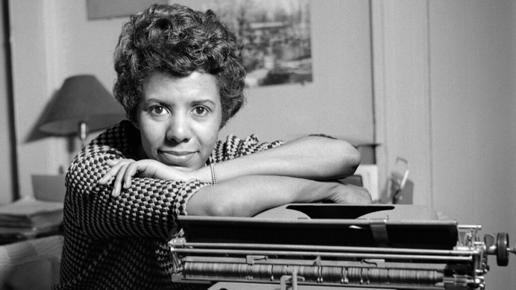 Lorraine Hansberry rests her chin on her crossed arms atop a typewriter. She smiles gently and looks into the camera, seated at a desk.