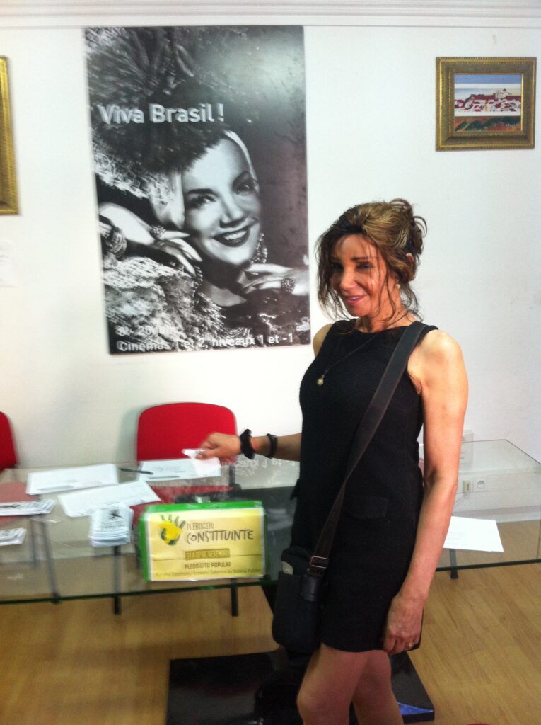 Camille Cabral smiling at a voting station indoors, standing in front of a large black-and-white poster that says “Viva Brasil!” while holding a voting slip.