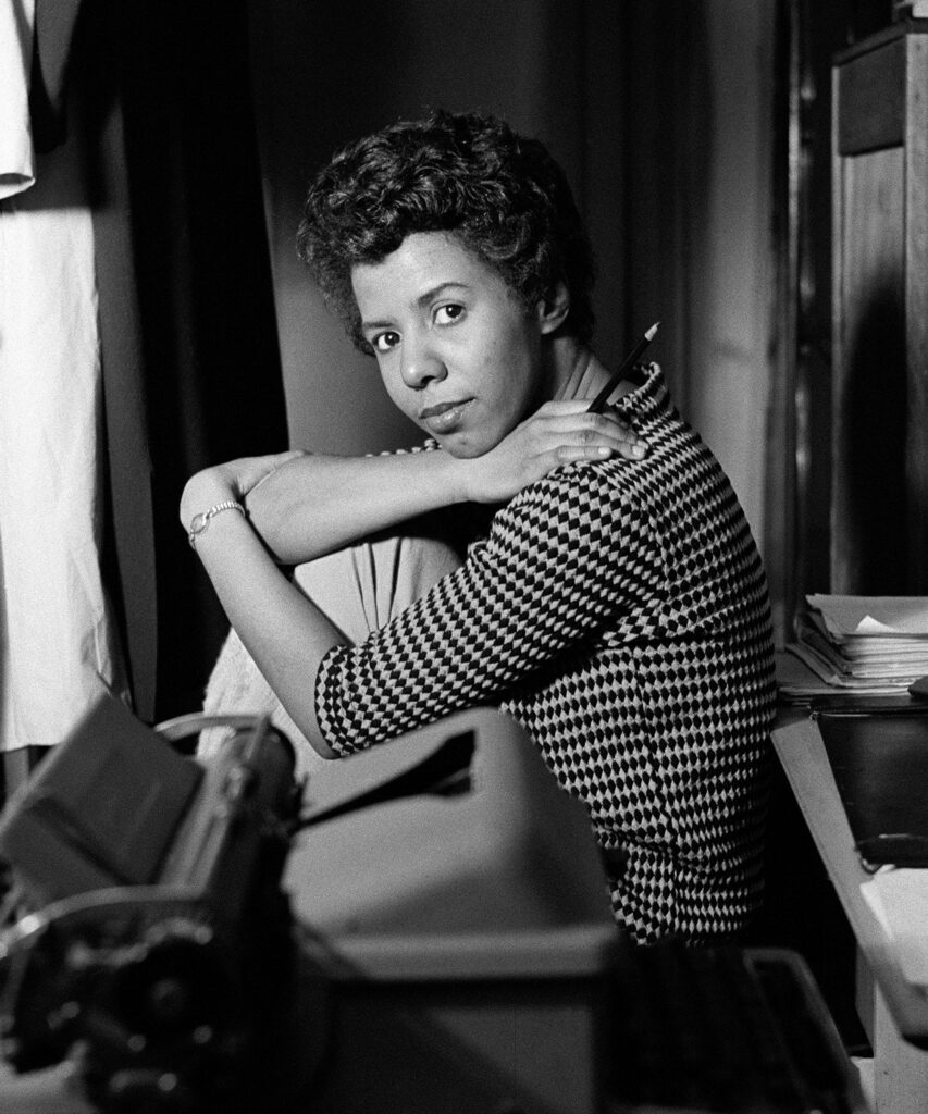 Lorraine Hansberry sits cross-legged near a typewriter, holding a pencil and gazing directly at the viewer with intensity.