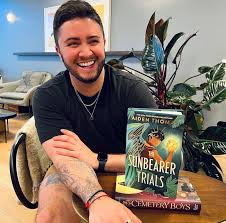 Aiden Thomas sits at a table grinning, with his books The Sunbearer Trials and Cemetery Boys stacked in front of him. A plant and cozy interior are visible in the background.