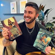 Aiden Thomas holds up copies of Cemetery Boys and The Sunbearer Trials, smiling widely while seated indoors with framed art on the wall behind him.