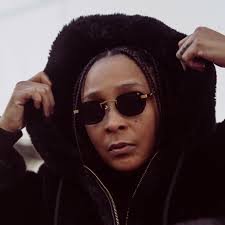 Felicia Pearson wearing dark sunglasses and a black hoodie with the hood pulled up, holding the edges with both hands. Her expression is cool and self-assured.