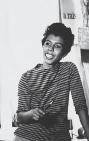 Smiling Lorraine Hansberry stands casually with a pencil in hand, in front of a poster that partially shows the words “A Raisin…”