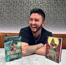Aiden Thomas laughs while sitting behind copies of The Sunbearer Trials and Cemetery Boys, with a tattooed arm resting on a marble table. A textured wallpaper is in the background.