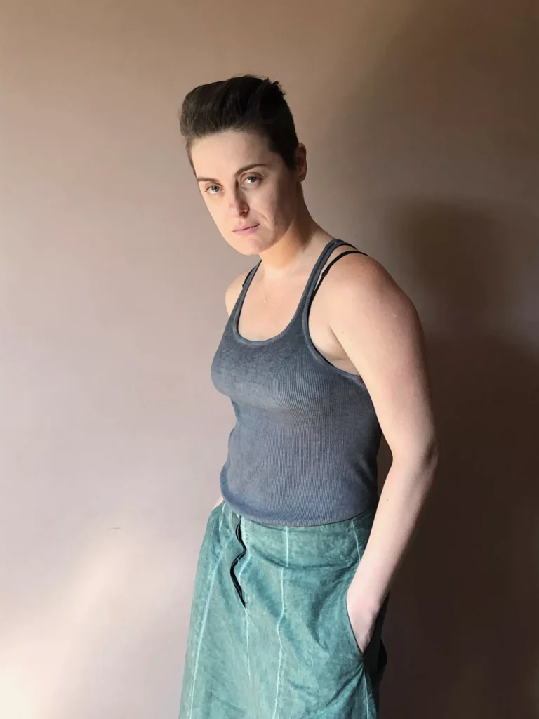 Alessandra Maestrini stands against a warm-toned wall in a gray tank top and teal skirt, gazing directly at the camera with a serious expression.