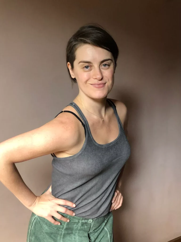 Alessandra Maestrini poses with hands on hips in a gray tank top and teal skirt, smiling confidently in front of a warm-toned background.