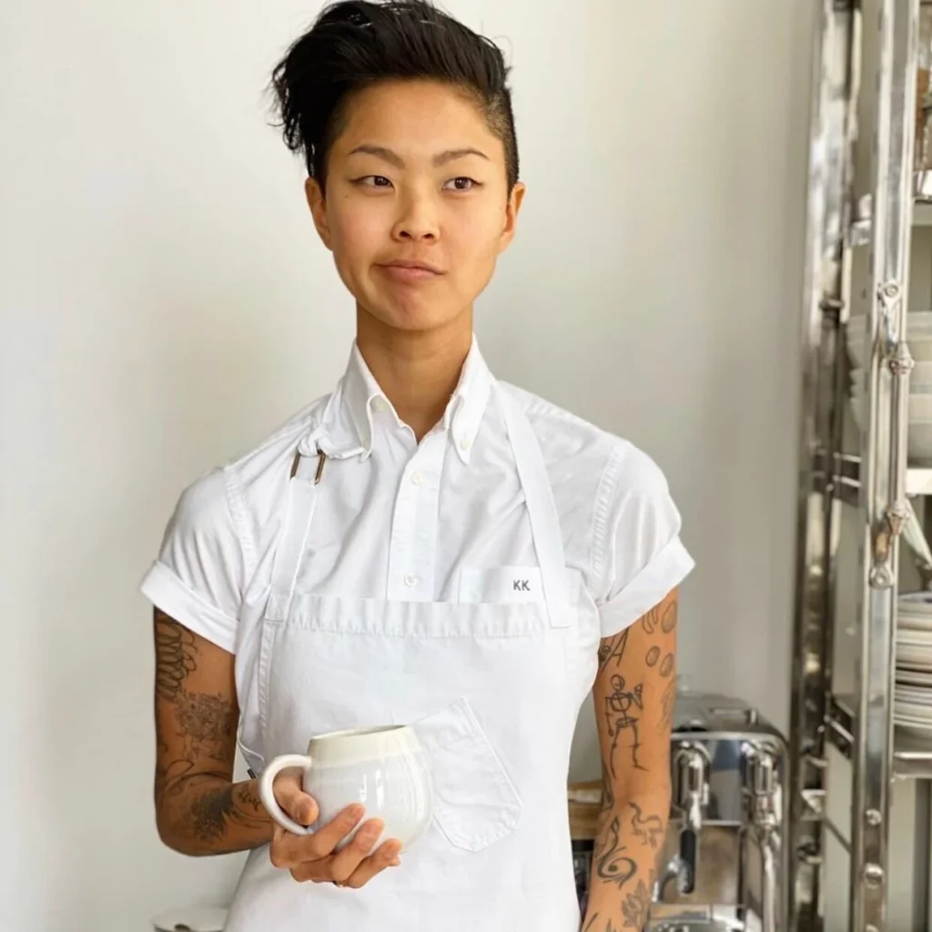 Kristen Kish in a crisp white short-sleeved shirt and apron labeled “KK,” holding a white mug, standing in front of metal shelving with a goofy expression.