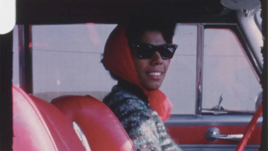 Lorraine Hansberry wears sunglasses and a red scarf while sitting in the driver’s seat of a vintage car, smiling at the camera.