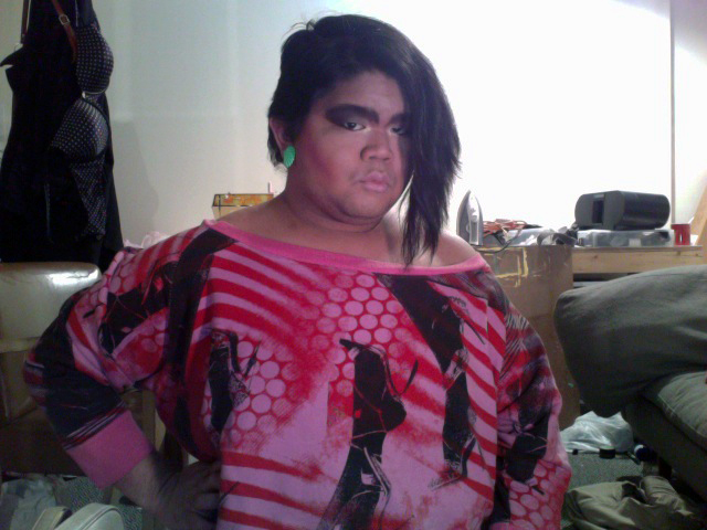 A bold selfie of CallOutQueen in full glam: exaggerated black eye makeup, neon green earrings, and a red-and-black off-shoulder graphic top. She glares into the camera with fierce defiance.