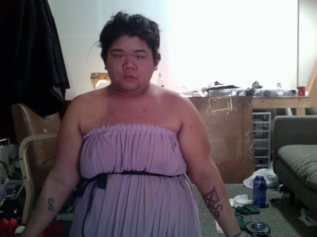 CallOutQueen wears a strapless lavender dress and stands with her arms relaxed at her sides, tattoos visible. The space around her is cluttered with supplies and lived-in textures, reinforcing her raw, unfiltered aesthetic.