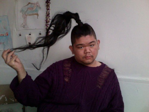 CallOutQueen sits in a purple knit sweater with torn shoulder detailing, holding up a long ponytail extension. She gazes into the camera with dry humor and unimpressed sass.