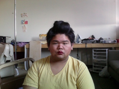 In a yellow t-shirt and bright lipstick, CallOutQueen sits with styled hair in a high pompadour. The studio behind her includes an ironing board, pinned notes, and the everyday mess of creation. She stares forward with sharp awareness.