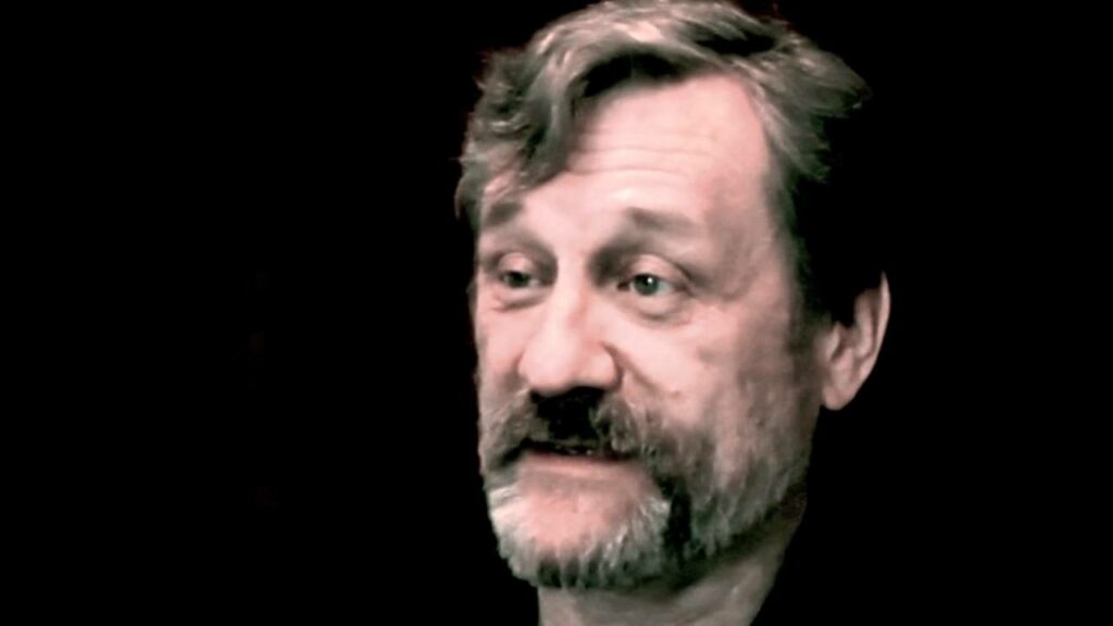 A close-up video still of Michael Bronski with a beard and mustache, speaking in front of a dark background. He appears mid-sentence, lit from the front.