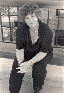 Joan Nestle sitting on a city rooftop, dressed in a dark shirt and pants, hands clasped, with short curly hair and a thoughtful gaze.