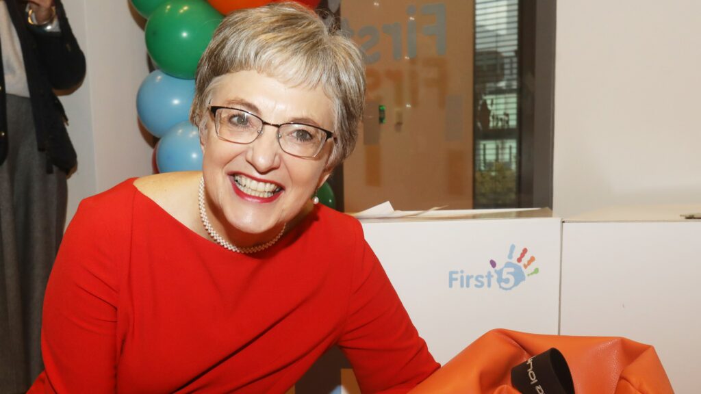 Katherine Zappone smiling in a red dress at a First5 early childhood event, with colorful balloons and branding visible in the background.