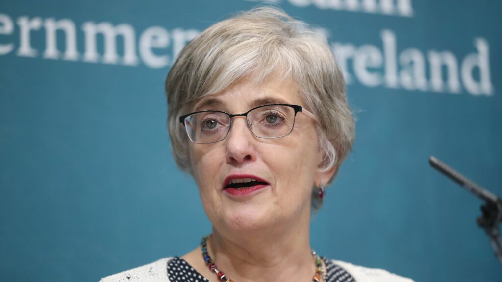Katherine Zappone speaking at a government press event, wearing glasses and a white top with black polka dots, standing in front of a teal backdrop with "Government of Ireland" partially visible.