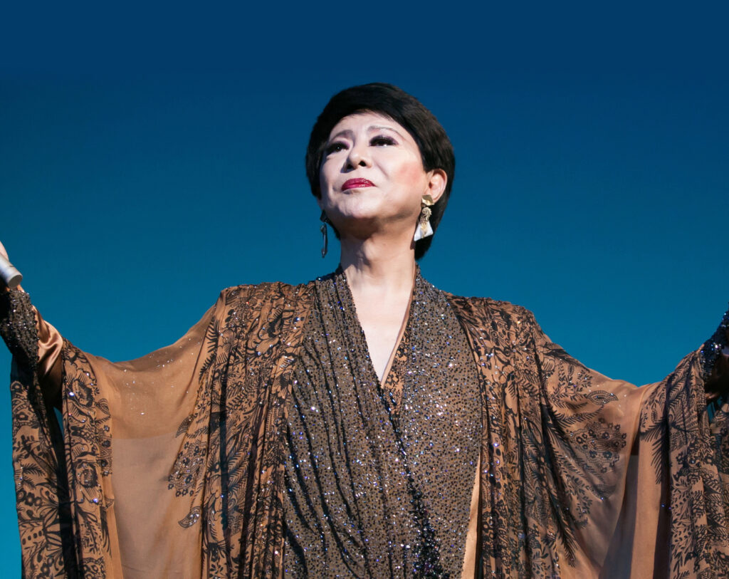 Kenichi Mikawa performs under stage lights wearing a luxurious bronze and black sequined robe with intricate patterns. His arms are outstretched and his face lifted toward the light, radiating a commanding and theatrical presence.