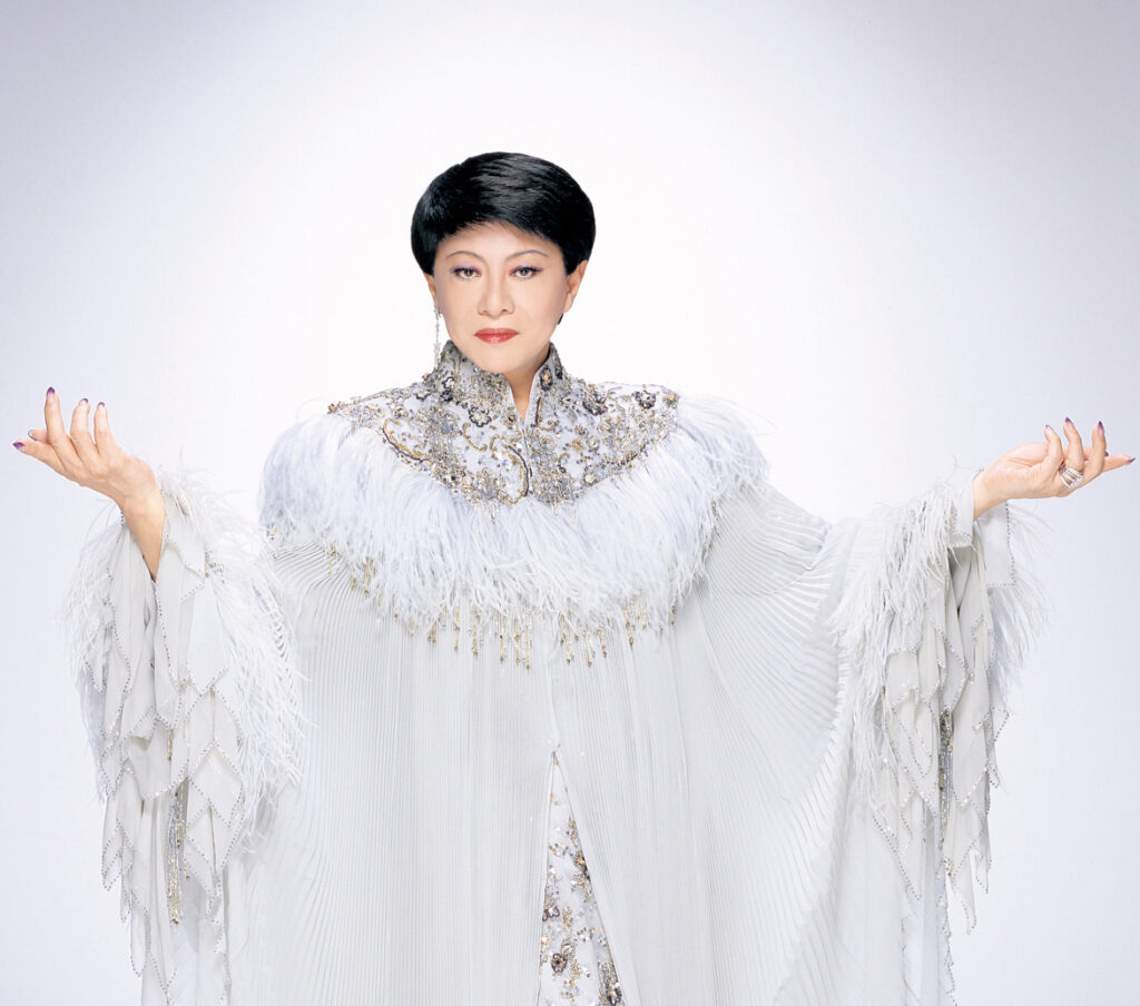 Kenichi Mikawa appears in a regal white outfit adorned with feathers and gold embroidery. His arms are raised outward in a dramatic gesture against a white backdrop, creating a divine, almost angelic silhouette.