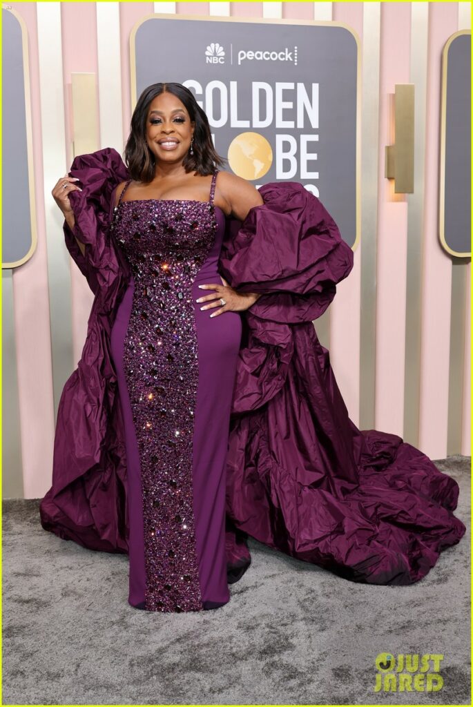 Nash-Betts radiates glamour in a jewel-embellished deep purple gown with dramatic puffed sleeves and a long train, posing on the red carpet at the Golden Globe Awards with a confident smile.
