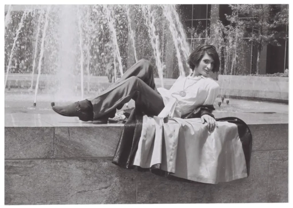 A black-and-white image of a young Sylvia Rivera reclining confidently on the edge of a fountain in a white blouse, flared pants, and leather jacket, exuding carefree charm and revolutionary spirit.