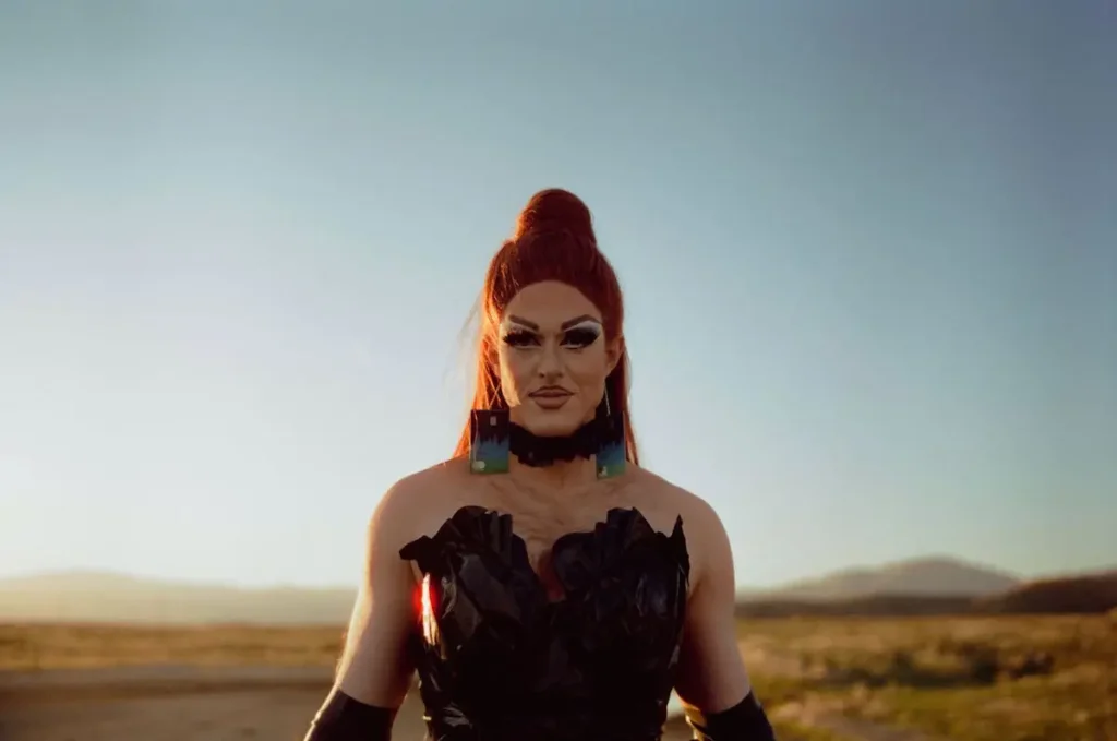 Pattie Gonia stands in an open field wearing a dramatic black dress made of recycled materials with matching black gloves and oversized earrings, lit by the golden hour sun.