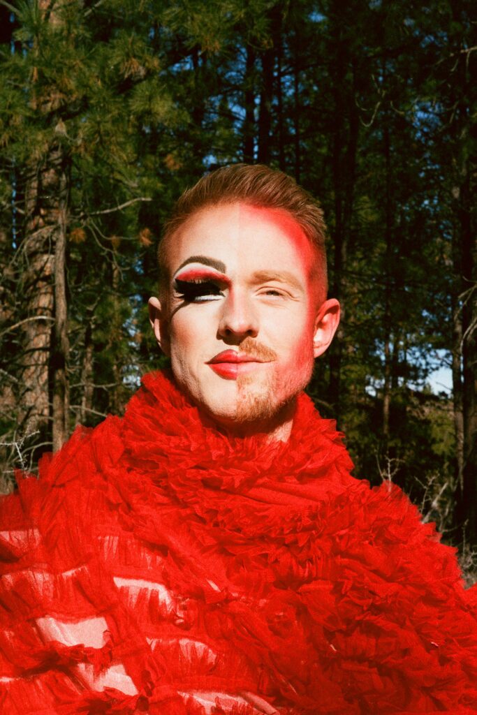 Close-up of Wyn Wiley’s face divided into two halves: the left side in full drag makeup with heavy lashes and red lipstick, the right side bare-faced. He wears a vibrant red ruffled outfit, standing in front of pine trees.