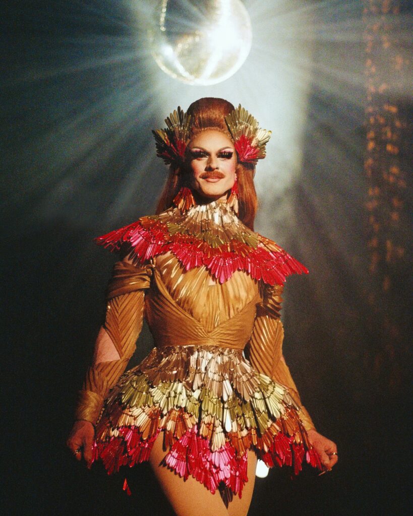 Pattie Gonia stands confidently under a glowing disco ball, wearing a structured drag outfit made of metallic gold, red, and silver feather-like pieces that resemble a bird's plumage. Her makeup is bold and her hair is styled in a high sculpted look.