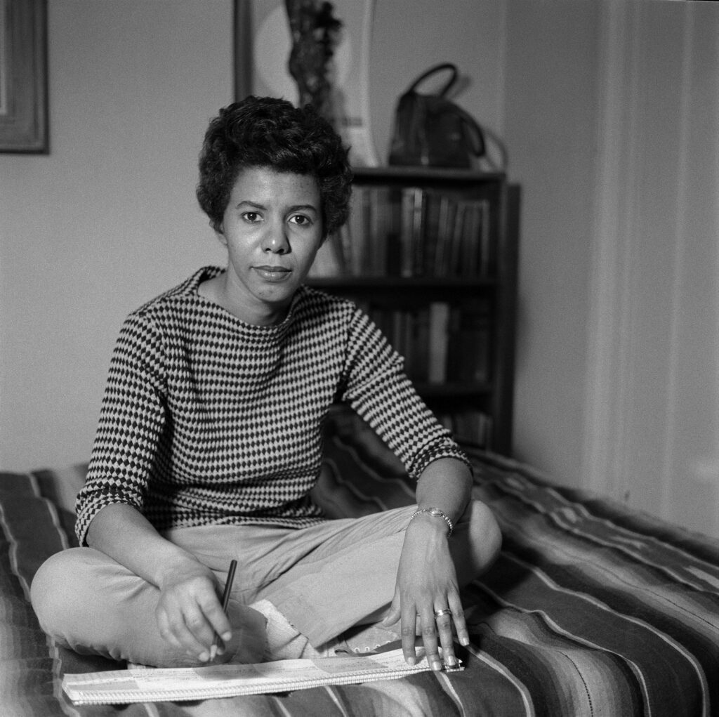 Lorraine Hansberry sits cross-legged on a bed with a striped blanket, pencil in hand, writing on a legal pad with bookshelves in the background.
