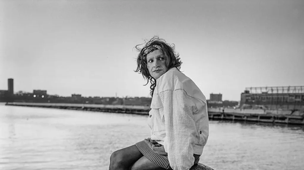Sylvia Rivera on Pier – Hudson River
Black-and-white image of Sylvia Rivera sitting on a dock by the water wearing a white jacket, skirt, and headband. Her expression is serious and thoughtful, with the city skyline in the background.