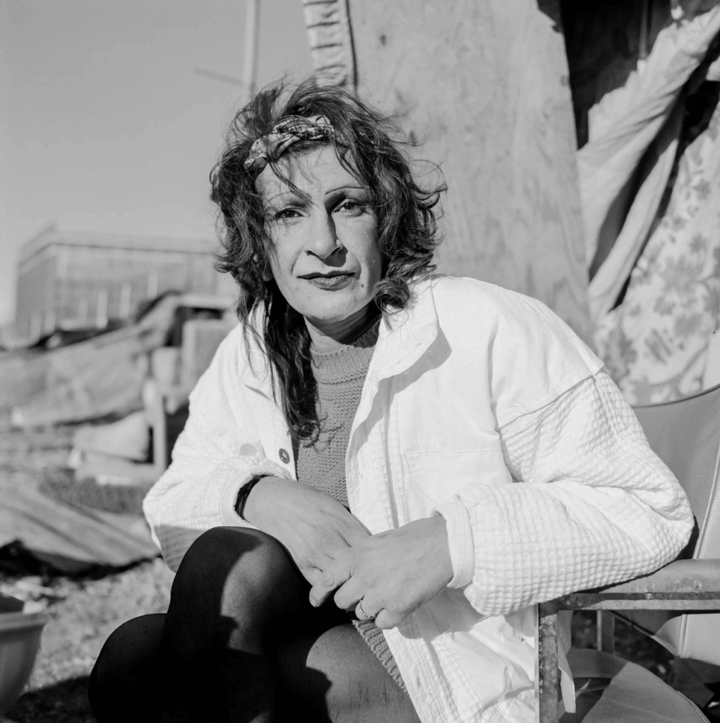 A striking outdoor black-and-white photo of Sylvia Rivera seated by the piers, wearing a textured white jacket and a headband. Her direct gaze speaks to her strength and history.