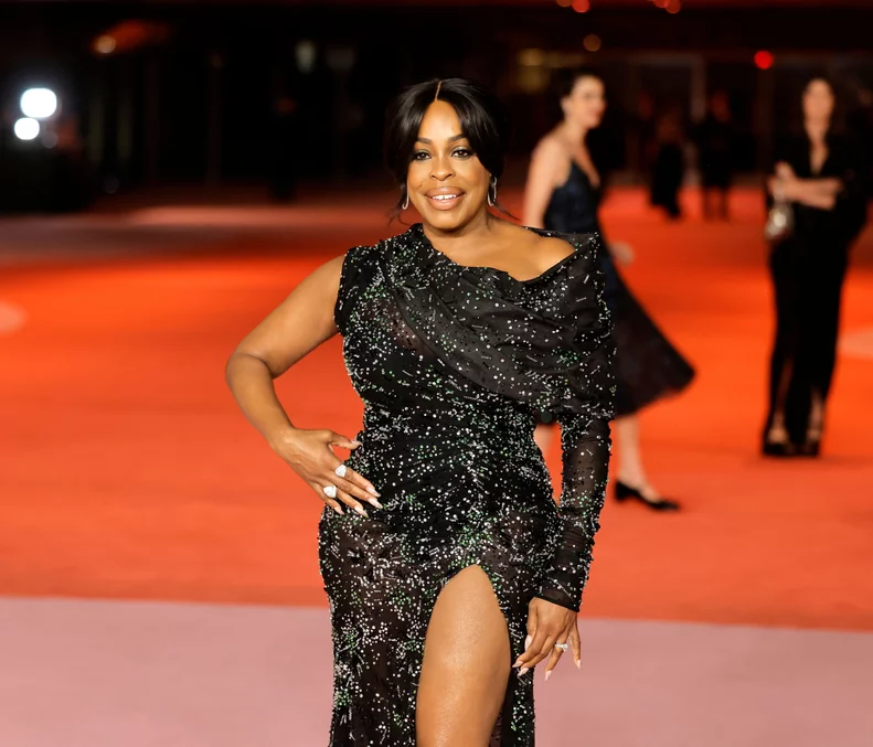 Niecy Nash-Betts stuns on the red carpet in a black, glittering one-shoulder gown with a high slit, her hand on her hip as she smiles with sleek hair and elegant jewelry.