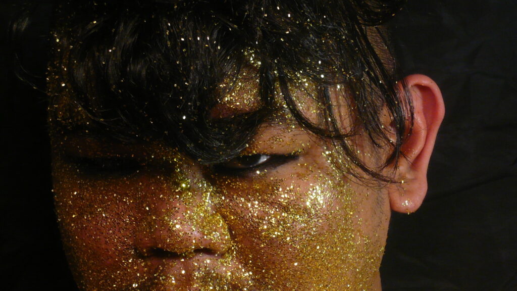 An extreme close-up of CallOutQueen’s face coated in shimmering gold glitter. Only one eye is visible beneath dark lashes and bangs, giving the image a celestial, sacred tone.