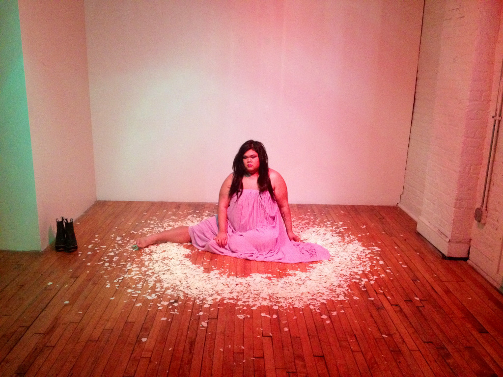 CallOutQueen sits on a polished wooden floor in a pink dress, surrounded by a wide circle of white flower petals. The lighting casts soft red and green glows, creating a ritual-like performance space where her body and presence are the centerpiece.