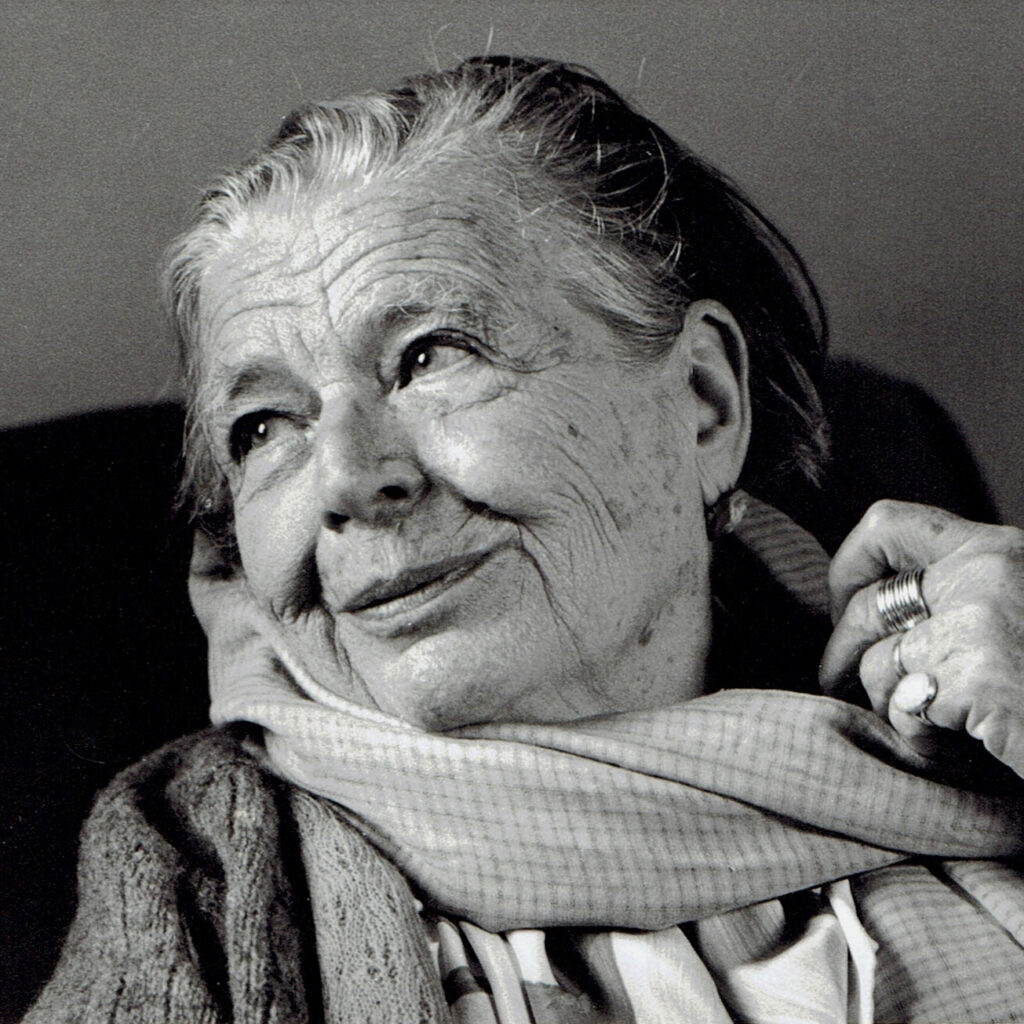 Elderly Marguerite Yourcenar with deep smile lines and a light scarf wrapped around her shoulders and neck, looking off to the side with a gentle expression.