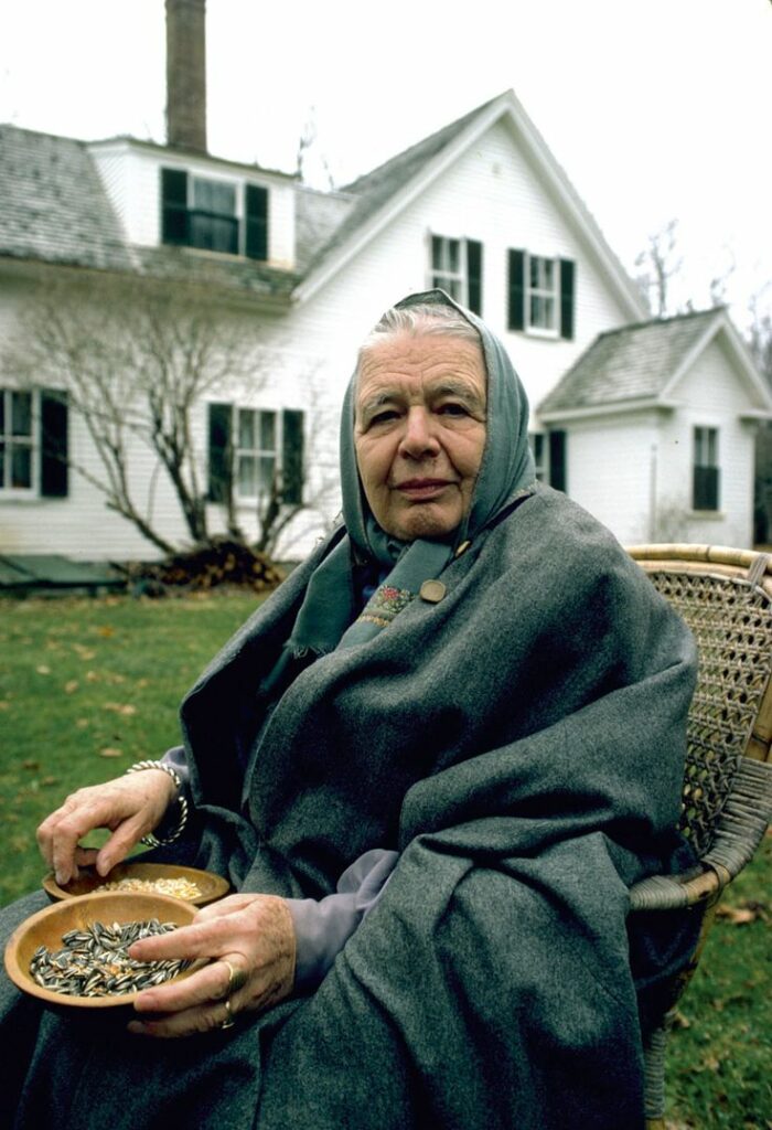 Marguerite Yourcenar sitting outdoors in a wicker chair, wrapped in a dark shawl with a scarf over her head, holding bowls of seeds in her lap, in front of her white house in Maine.