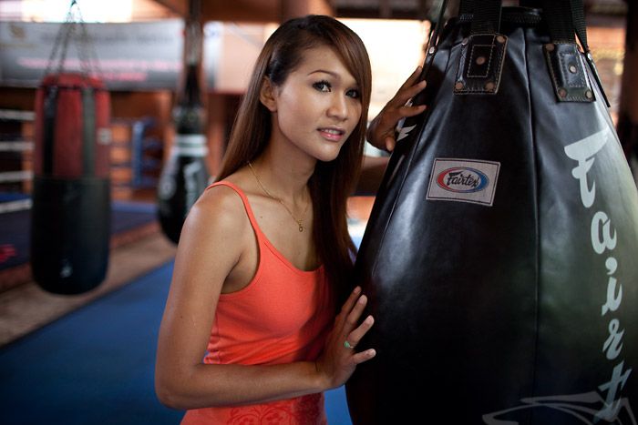 Parinya Charoenphol stands in a Muay Thai gym, wearing an orange tank top and posing next to a Fairtex punching bag, smiling warmly.