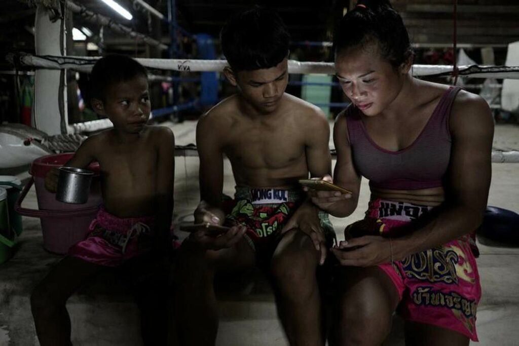 A young Parinya Charoenphol sits inside a Muay Thai ring with two other fighters, reviewing something on a phone. She wears a sports bra and Muay Thai shorts, while a young child with a metal cup sits nearby.
