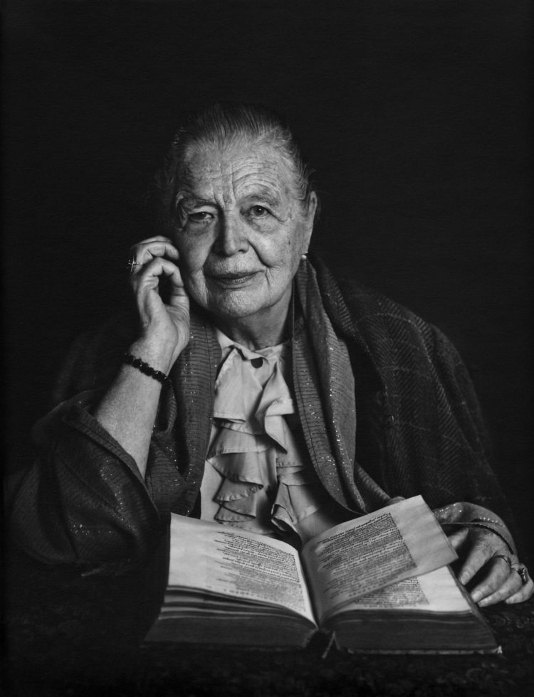 Striking formal portrait of Marguerite Yourcenar taken by Yousuf Karsh in 1987, seated with a large open book before her, looking directly at the camera with one hand resting near her face.