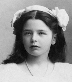 Vintage photo of Marguerite Yourcenar as a child with long dark hair, a white bow on each side, and a serious, contemplative expression.
