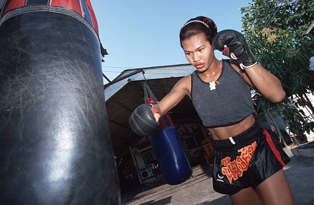 Parinya Charoenphol punches a heavy bag in an outdoor training area, dressed in a black tank top and Muay Thai shorts with her hair pulled back.