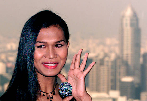 Parinya Charoenphol waves with a smile while holding a microphone, standing in front of a city skyline.