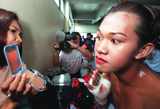 Backstage, Parinya Charoenphol applies face powder before a match, with hand wraps already on, while cameras and a makeup assistant surround her.