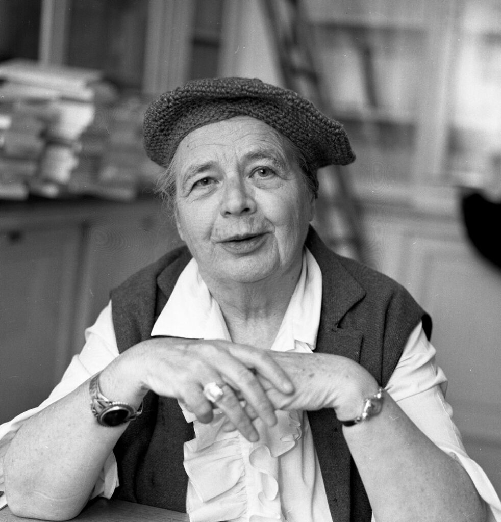 Photo of an older Marguerite Yourcenar seated at a table, smiling with her hands resting casually, wearing a knitted beret and sleeveless vest over a blouse.